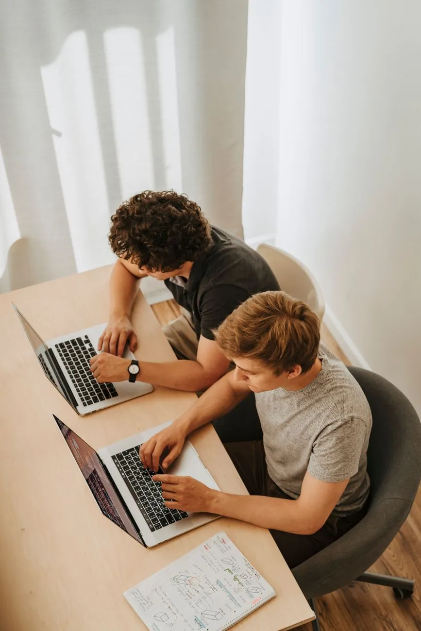 Two software engineers working side by side on laptops, notes sketched in a notebook