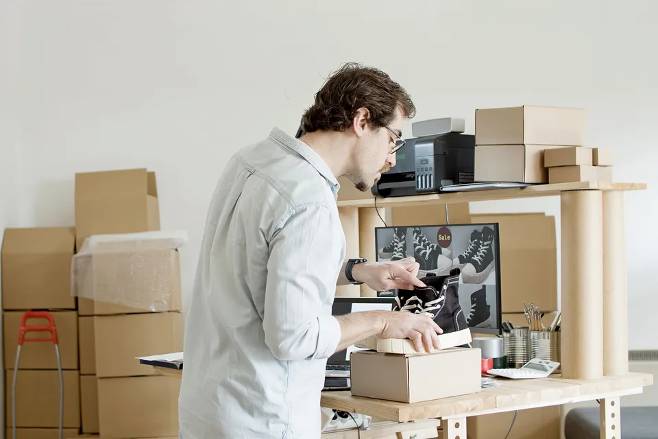 A store owner packing an online order with product photos visible on a monitor behind them