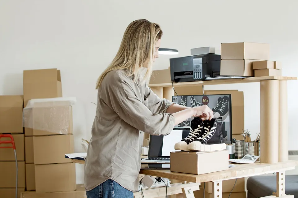 A store owner packing a pair of sneakers into a shipping box beside a laptop and a monitor showing a product catalog